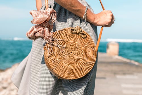 女人穿着灰色的裙子和圆形的棕色藤制斜挎包袋在木码头附近的水体
