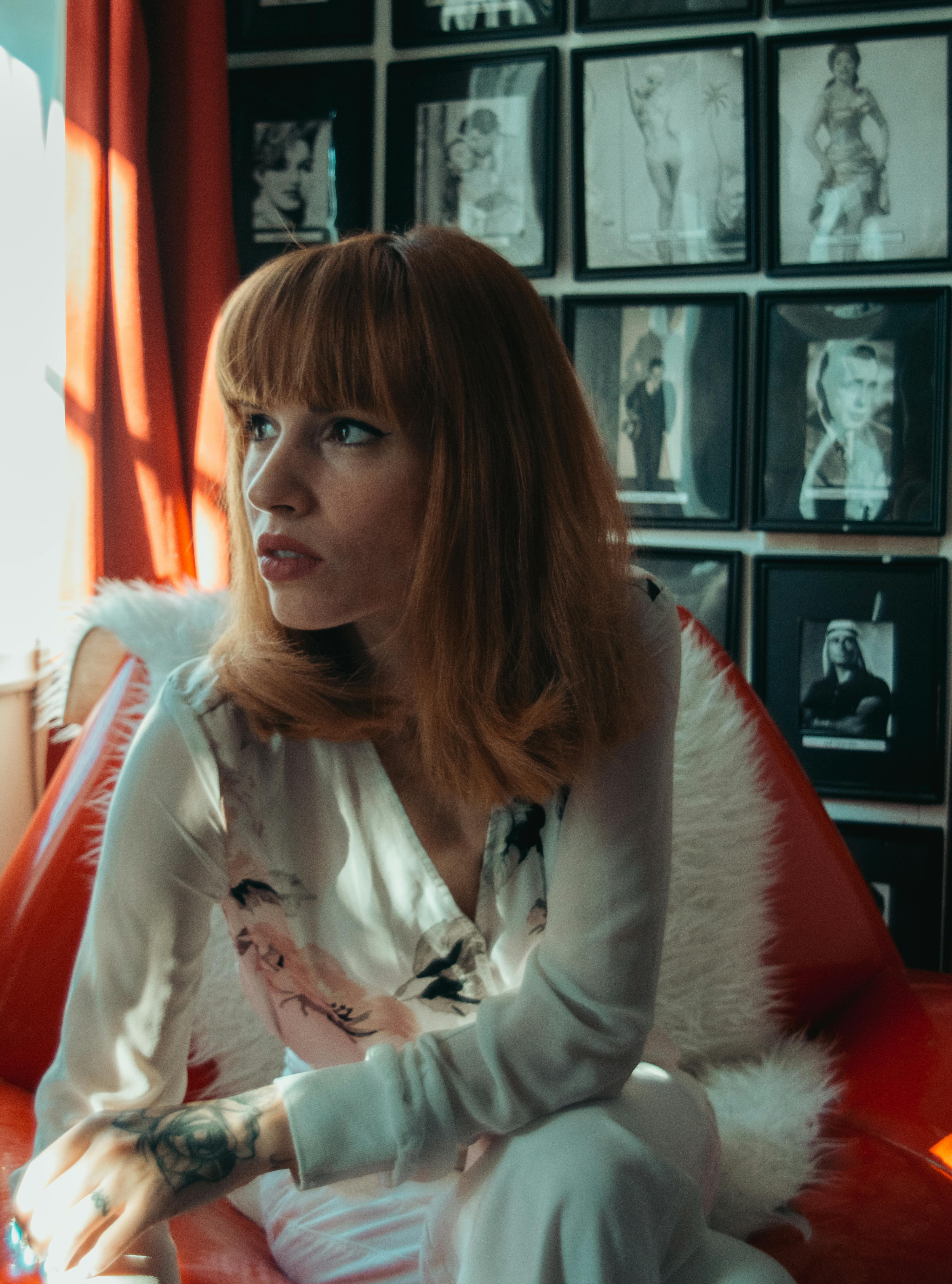 A thoughtful young woman in a stylish outfit sits on a retro couch surrounded by vintage decor.