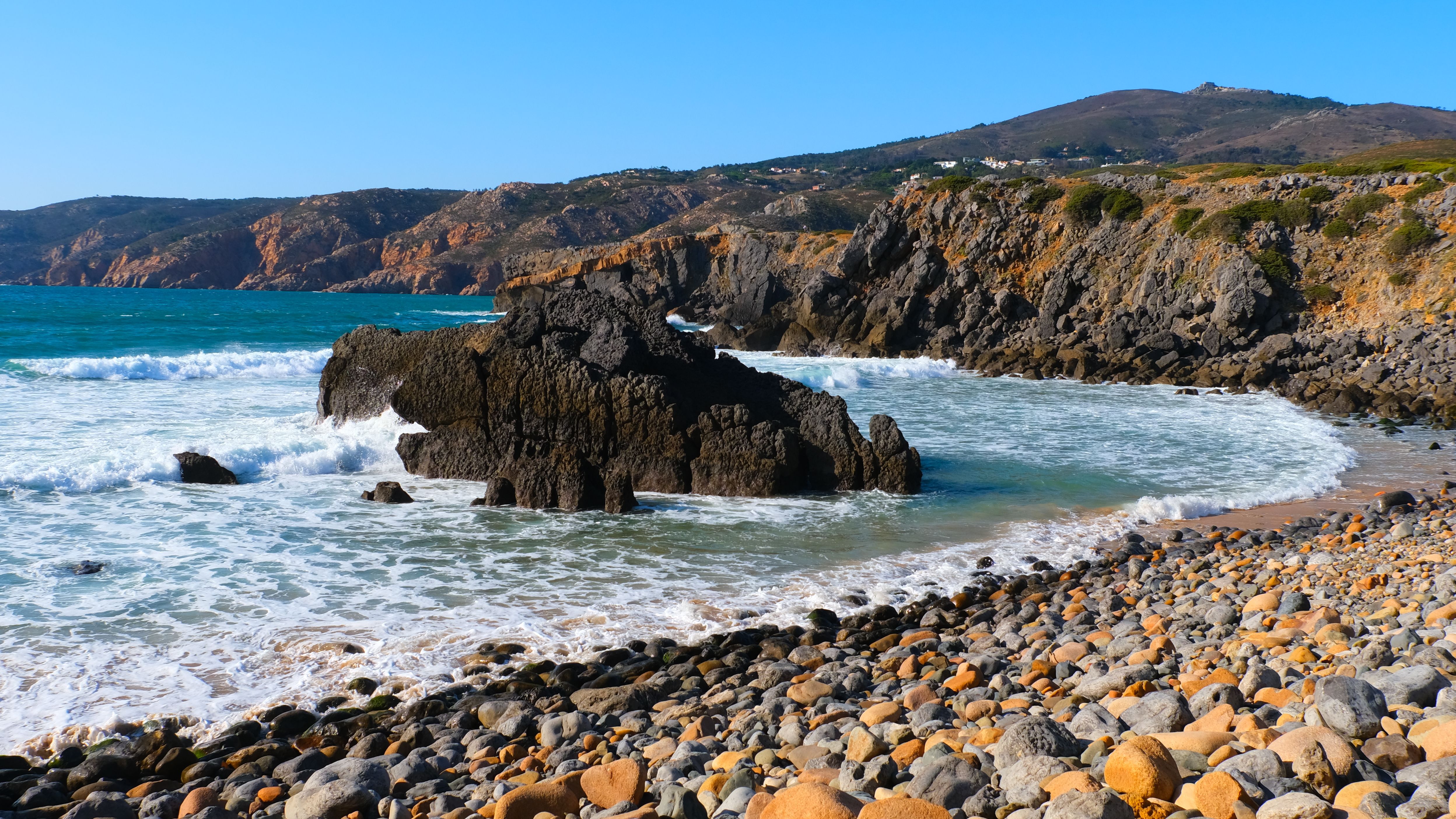 Scenic view of rocky beach with waves crashing on the shore under a clear blue sky.