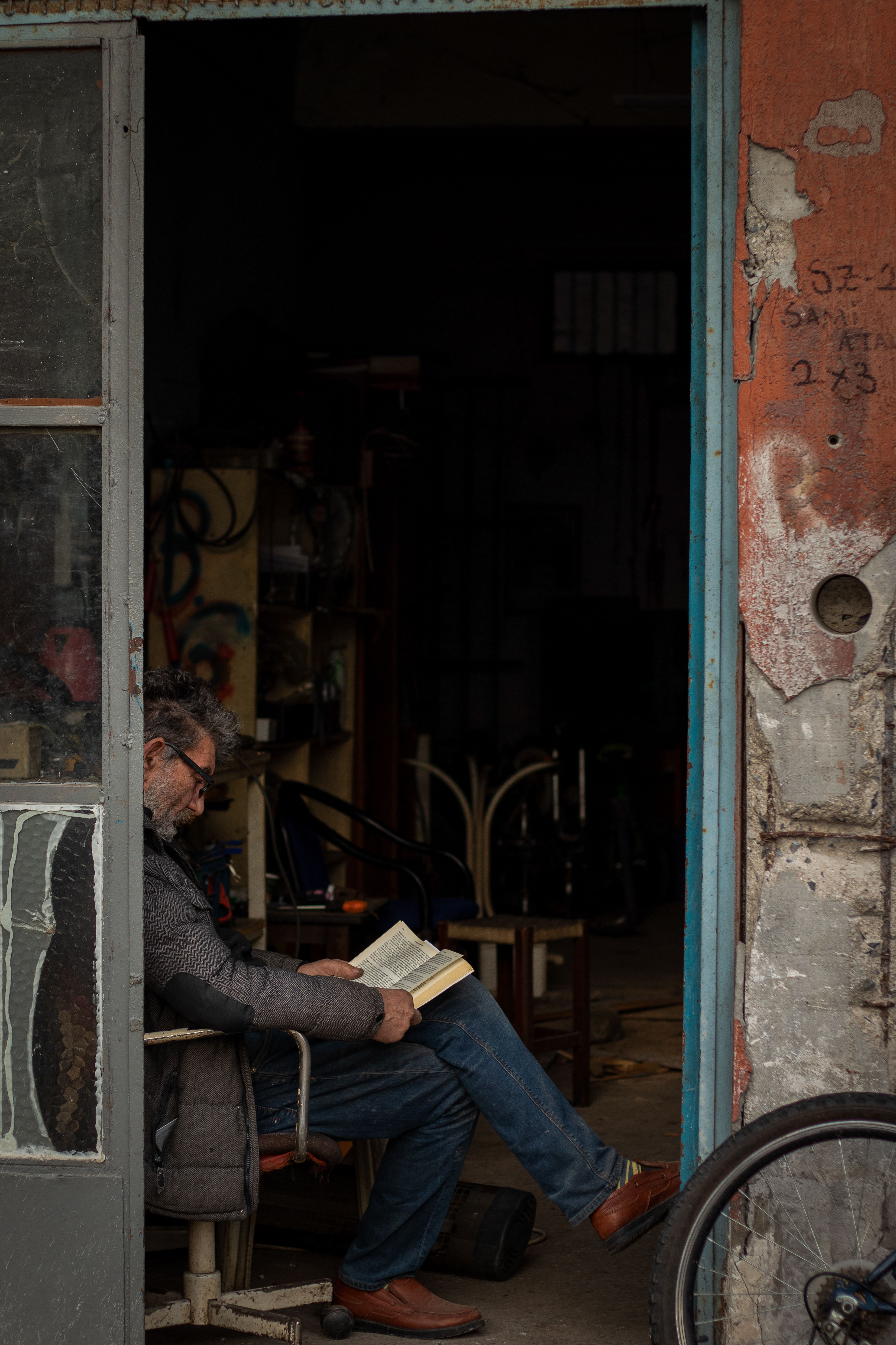 Senior man sitting on a chair reading a book inside a rustic workshop. Calm atmosphere.