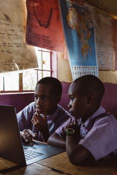 Two boys in school uniforms learning together on a laptop indoors with educational posters in the background.