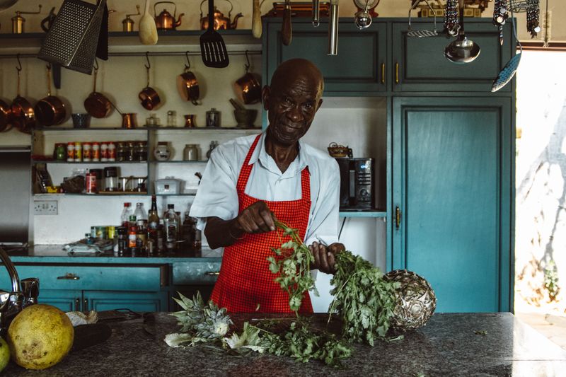 An older Black man in a white shirt and a red gingham apron stands at a kitchen counter stripping leaves from a bunch of fresh greens, with copper pots, jars and seasoning bottles on the shelves behind him.