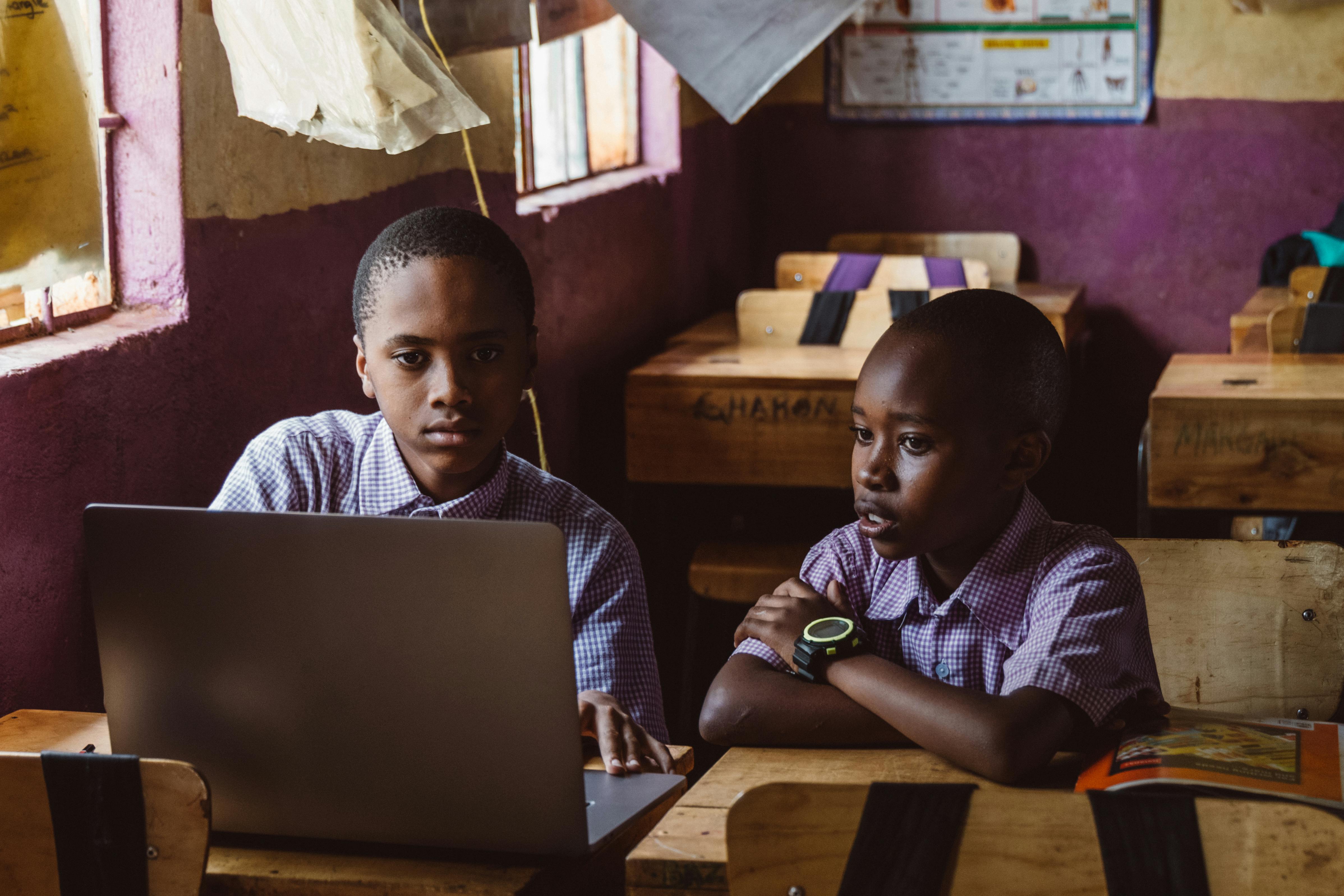 Two black boys in school uniforms focusing on a laptop, collaborating in a classroom setting.