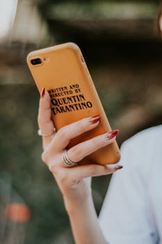 Elegant hand holding a smartphone with distinctive director-inspired phone case, showcasing nail art and rings.
