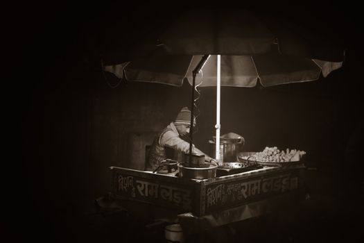 A grayscale photo of an Indian street vendor selling food at night under an umbrella.
