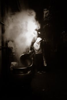 Monochrome street photography of a man in a white hoodie enveloped in cooking smoke in India.