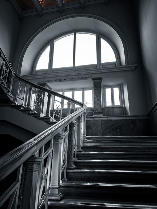 Free A grayscale photo of an elegant staircase and arched window in a historic building interior. Stock Photo