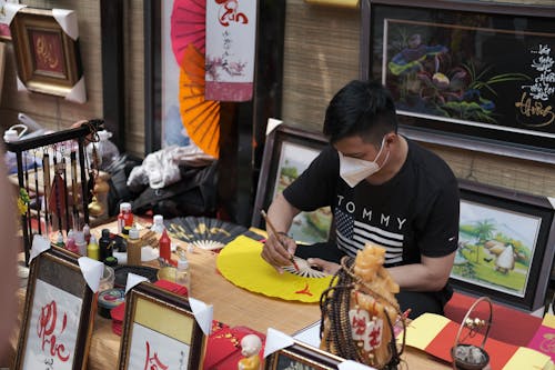 Free A Man in Black Shirt Wearing Face Mask while Painting on a Hand Fan Stock Photo