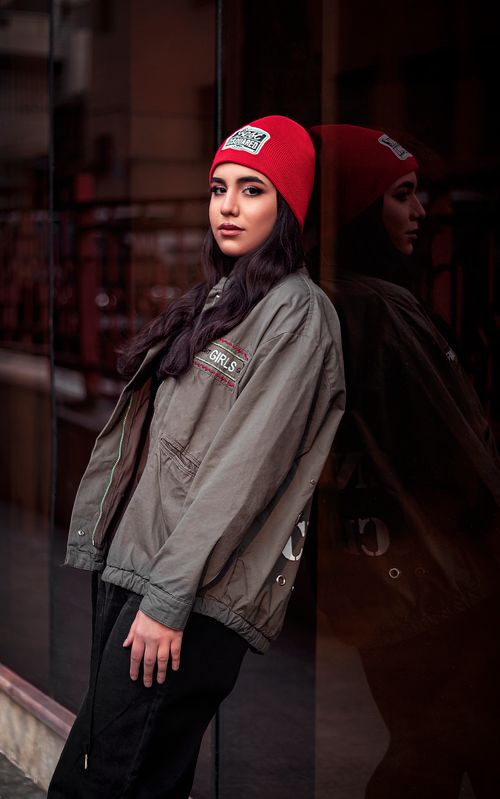 Free Stylish woman in Tehran wearing a red beanie and green jacket leaning against a reflective glass wall. Stock Photo