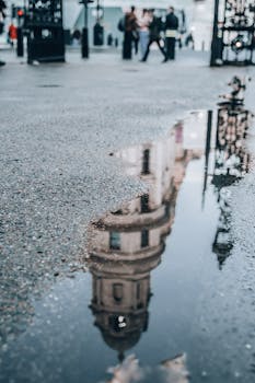 A dramatic reflection of a historic London building in a rain puddle with blurred pedestrians in the background.