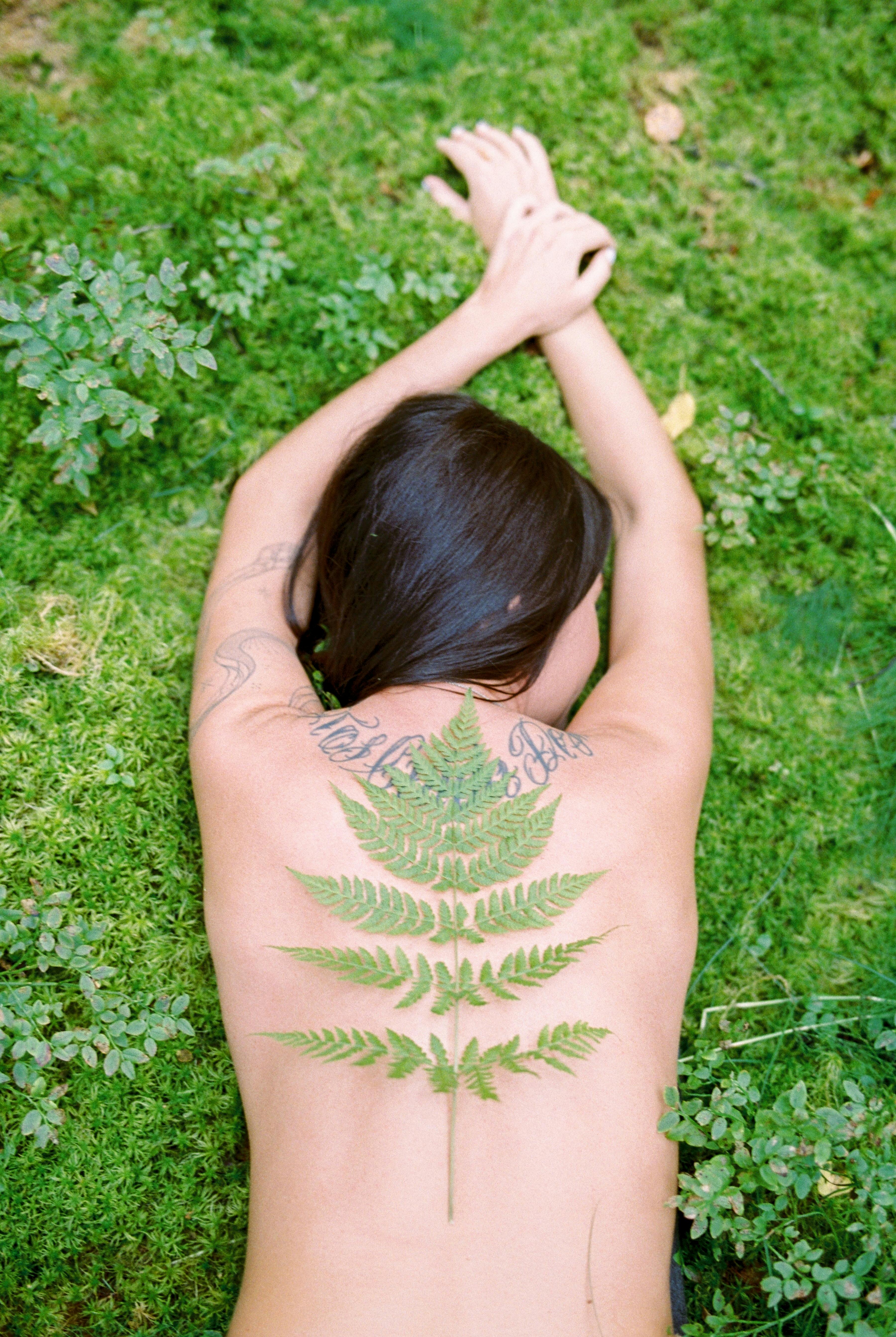 Nude woman lying on moss with a fern leaf placed on her back, showcasing natural harmony.