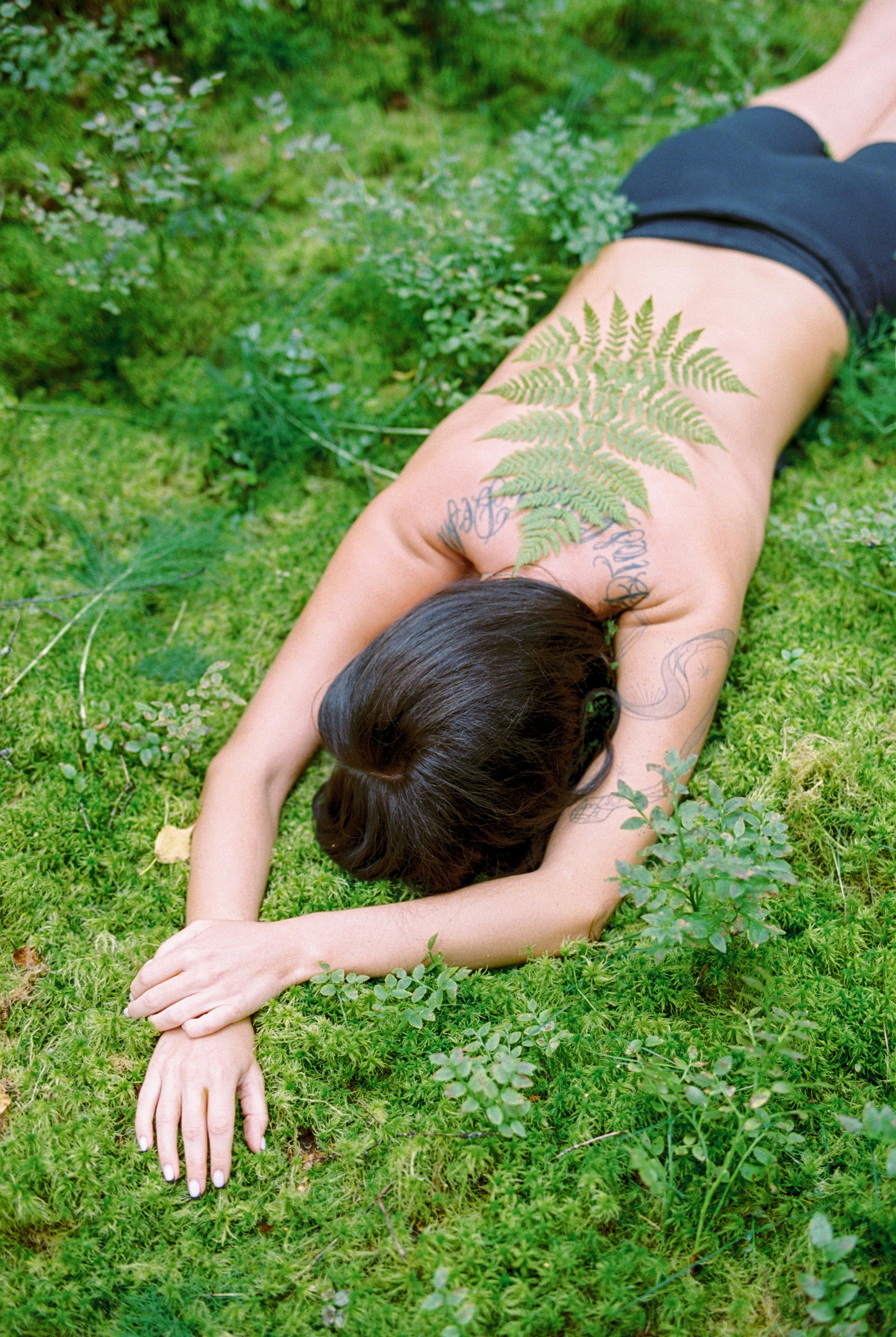 A topless woman with tattoos lies on a mossy field, blending with nature.