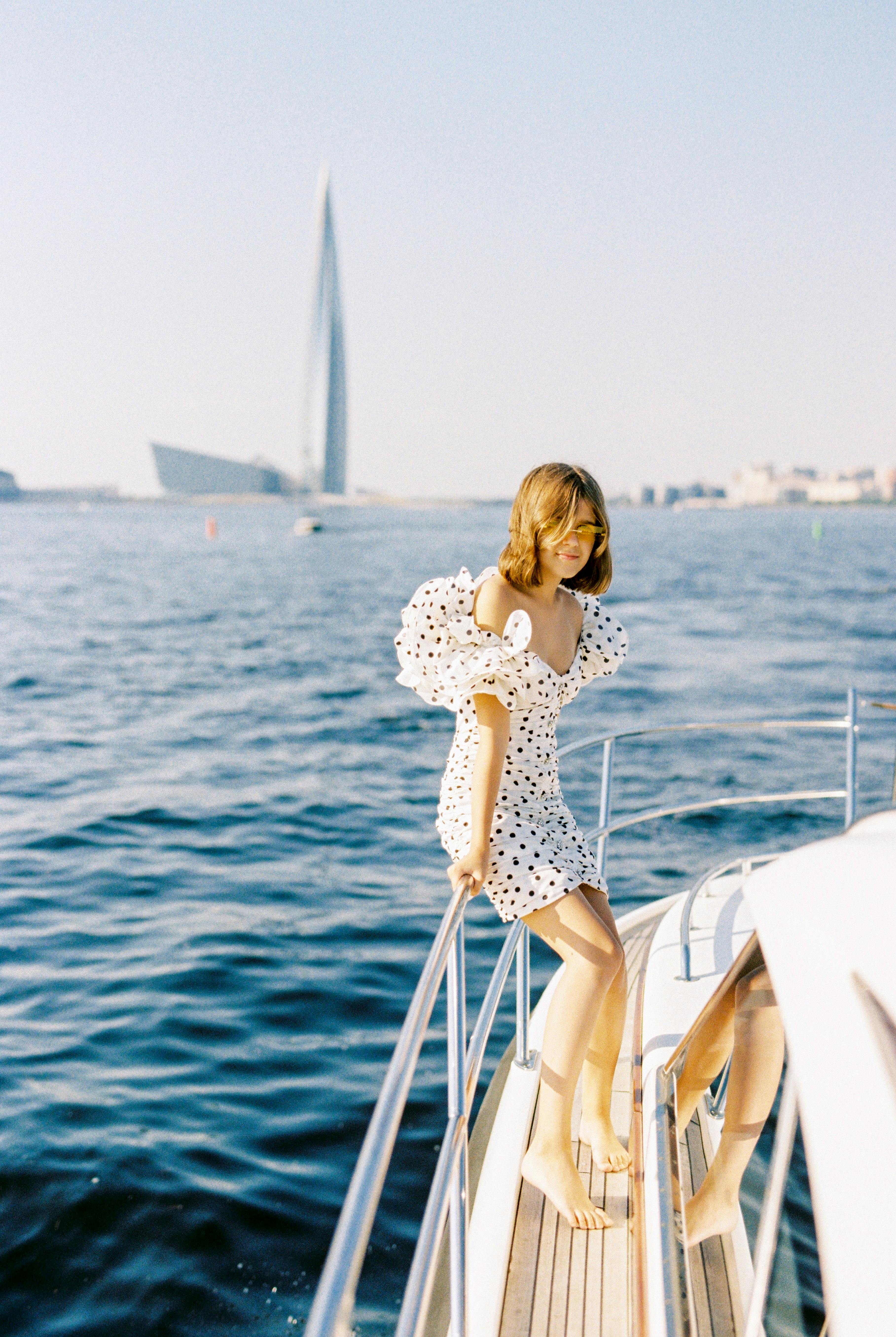 A woman in a polka dot dress stands on a boat deck with a city skyline in the background.