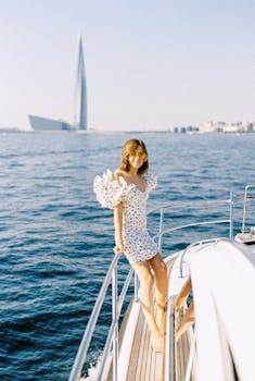 Elegant woman in a polka dot dress enjoys a sunny day on a yacht with modern cityscape in the background.