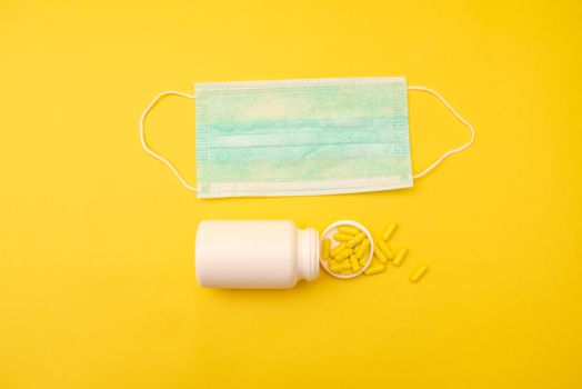 Close-up of a medical mask and yellow pills spilling from a bottle on a vibrant yellow background.