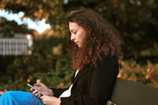 A young woman sits on a park bench in Warsaw, using her smartphone on a sunny day.