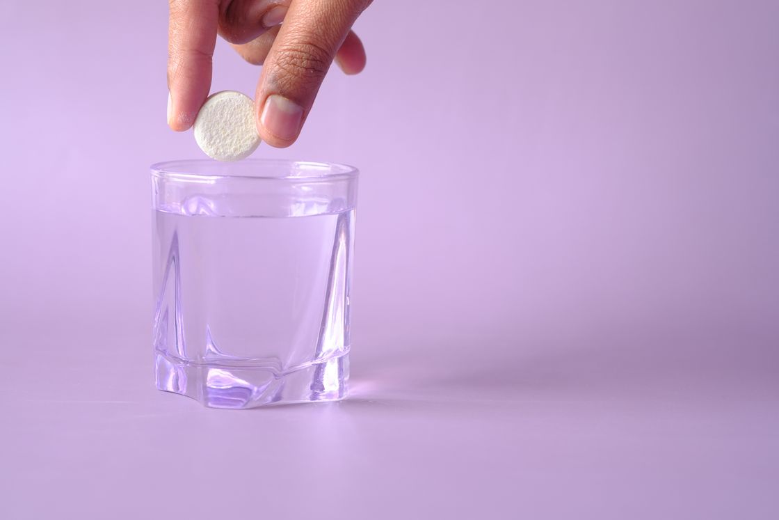 Hand dropping a white cleaning tablet into a glass of water on lilac