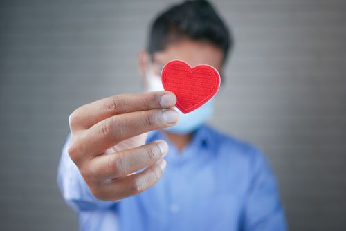 Free Close-up of a man with a mask holding a red heart symbol, symbolizing care and connection. Stock Photo