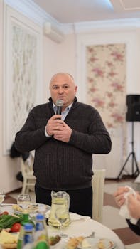 Bald Caucasian man holding a microphone while speaking at a formal indoor event.