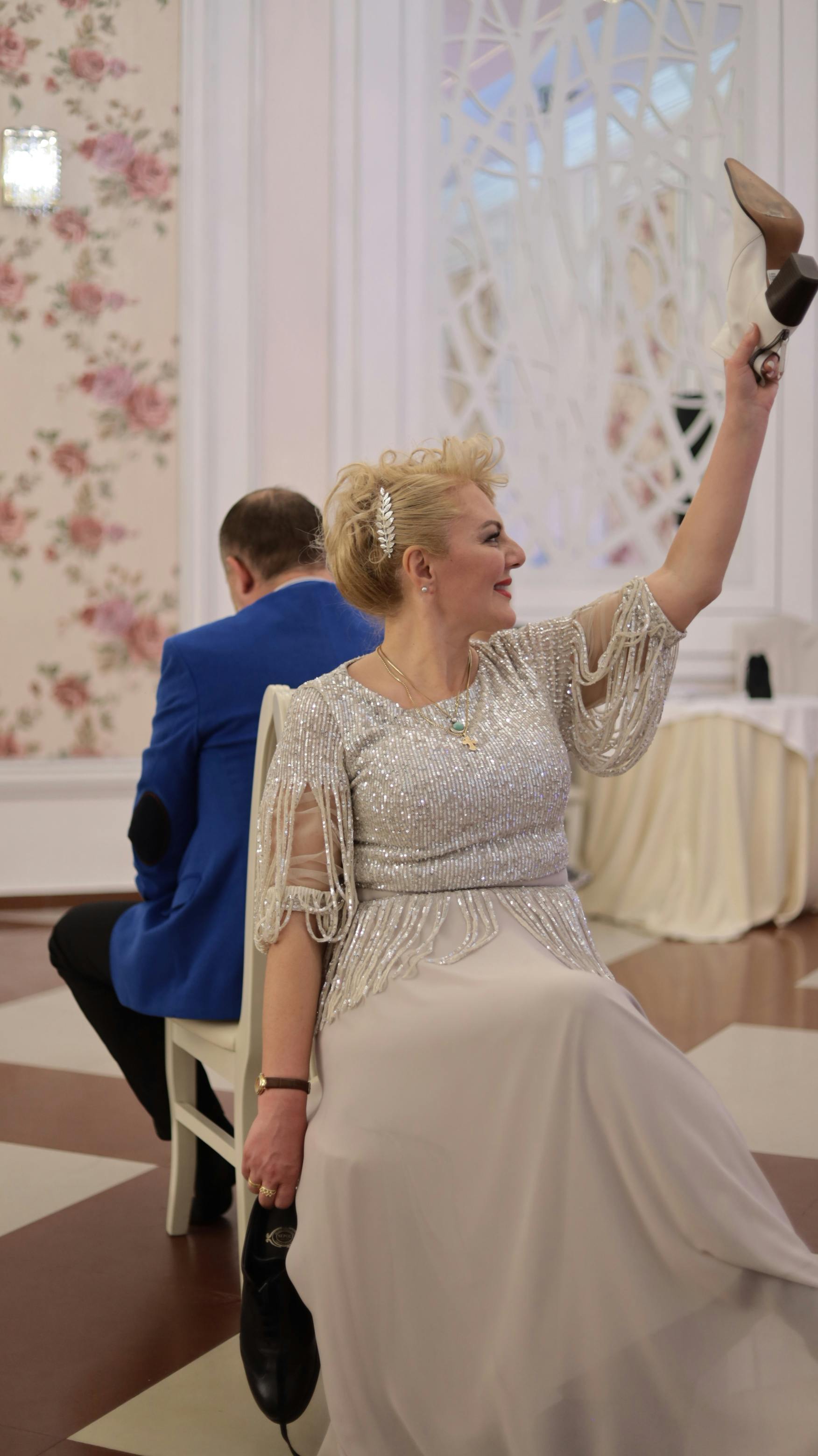 a woman in elegant white gown sitting on a chair while raising the shoe she is holding