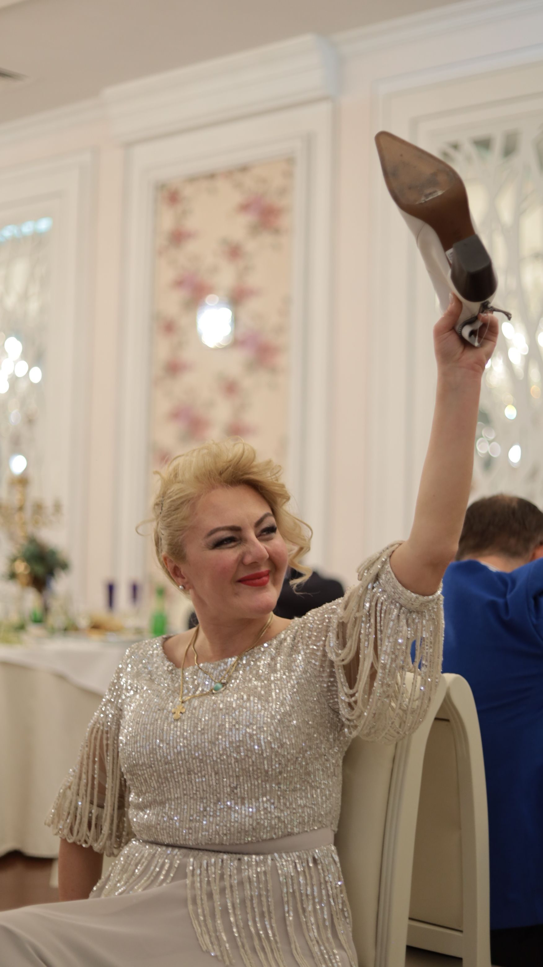 Joyful woman in a glittery dress, holding a shoe aloft during a party game indoors.