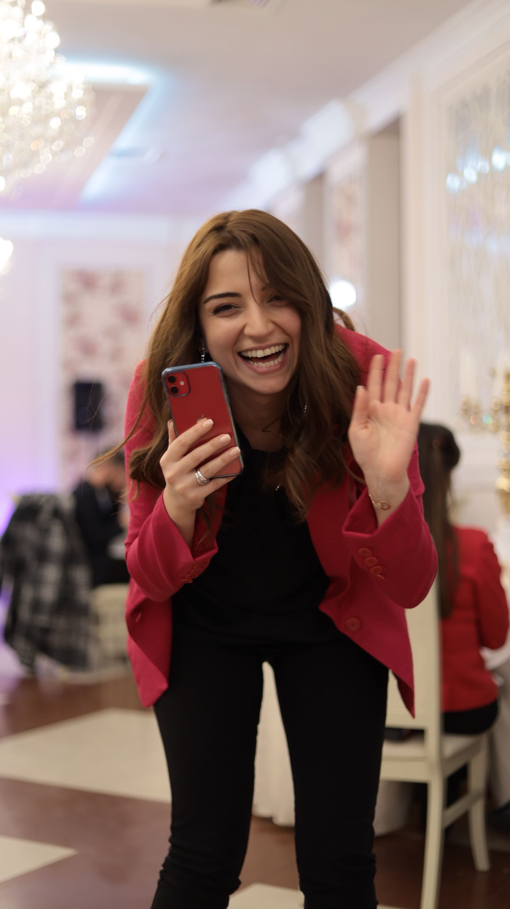 A cheerful woman in a red suit waves while holding a smartphone at a bright indoor event.