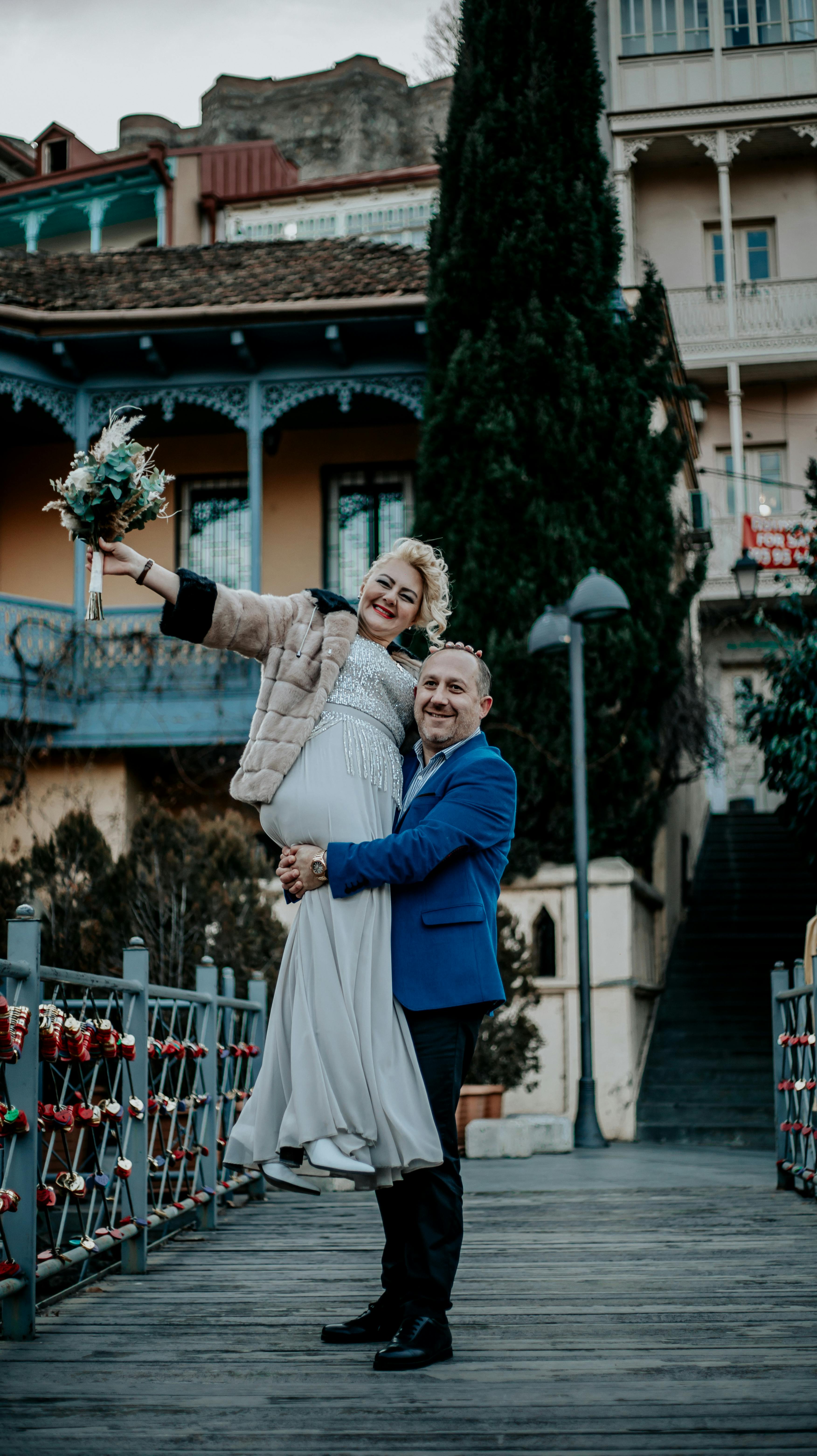 Smiling couple posing with bouquet on a charming pedestrian bridge outdoors.