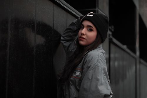 Free Stylish woman wearing gray jacket and beanie posing confidently against a black wall. Stock Photo