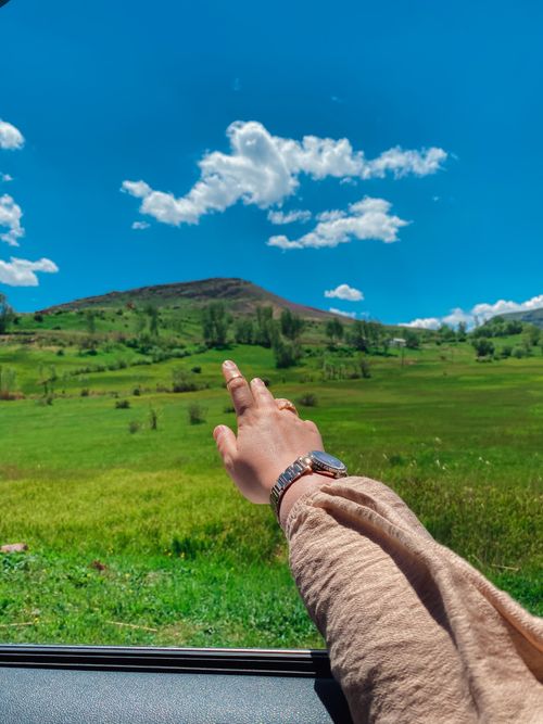 Free A hand reaching out from a car window towards a vibrant green landscape under a blue sky with clouds. Stock Photo