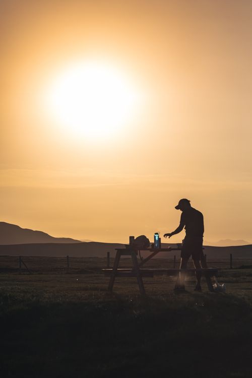 Free Silhouette of a person cooking at a picnic table during a stunning sunset, creating a serene outdoor atmosphere. Stock Photo