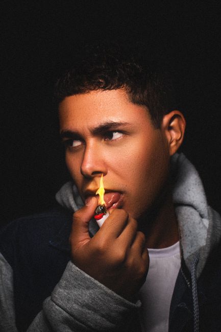 Young man in hoodie holding lighter to face, dramatic lighting on black background.