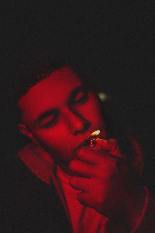 Free A man lights a cigarette with a lighter under dramatic red lighting, creating a moody atmosphere. Stock Photo