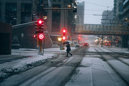 Person crossing a snowy street with umbrella at red traffic light during winter.