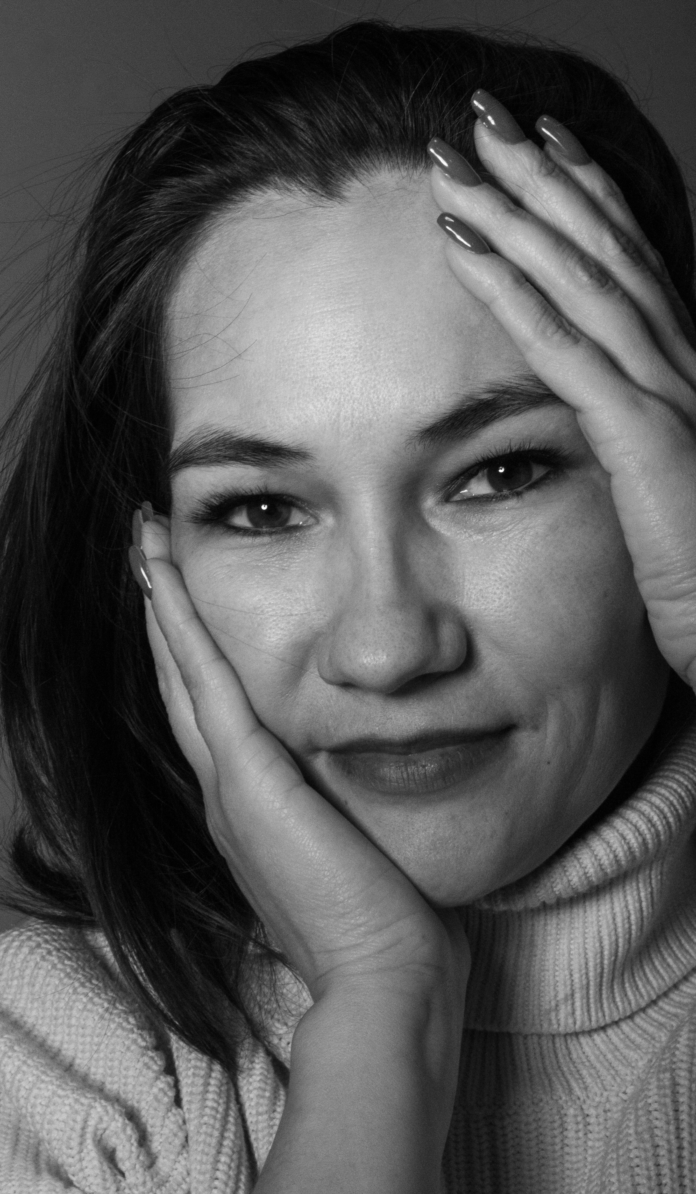 Grayscale close-up of a woman smiling, hands on face, wearing a turtleneck.