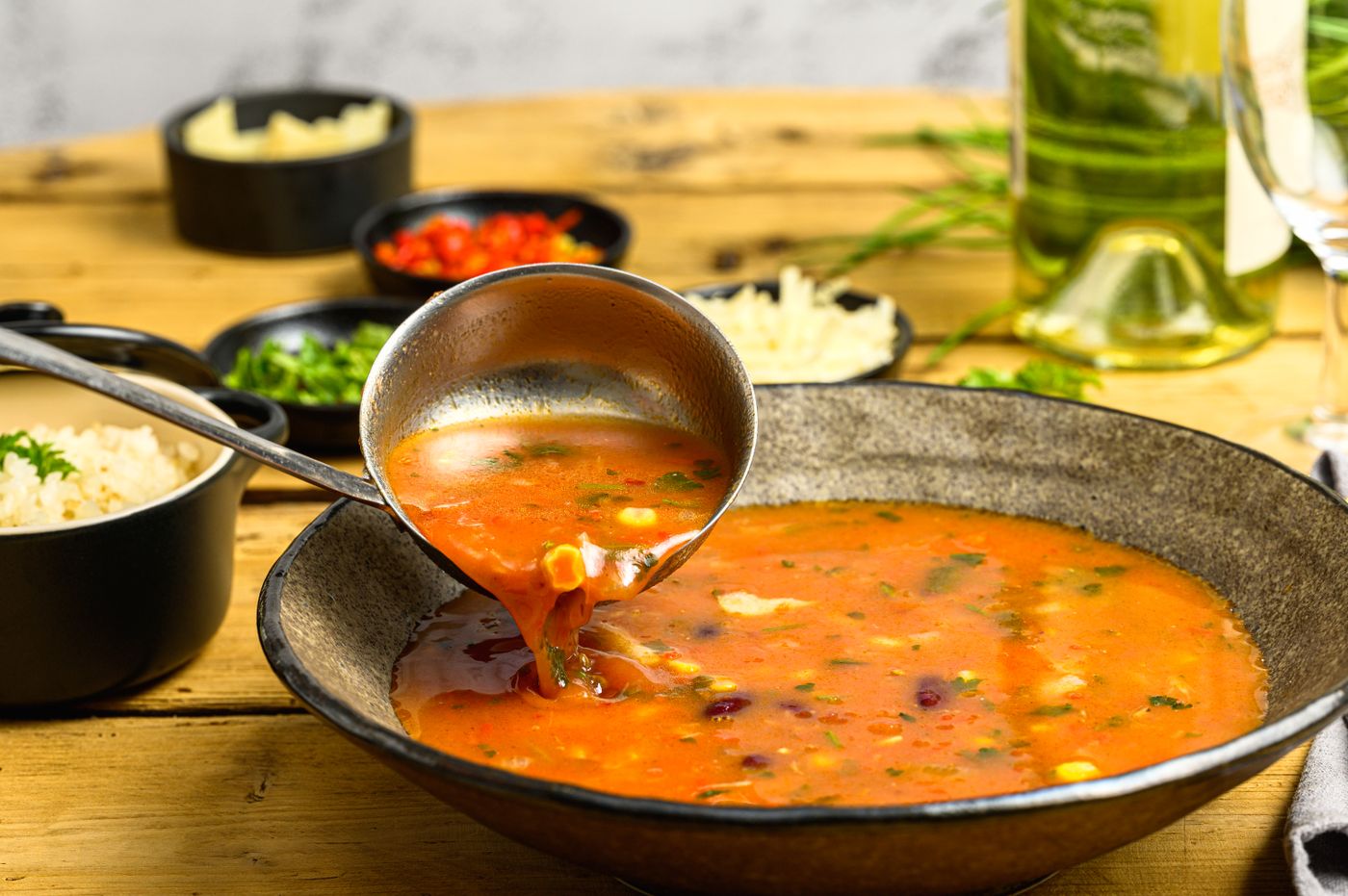 Close-up of a ladle serving vegetable soup in a rustic bowl with wine and side dishes.