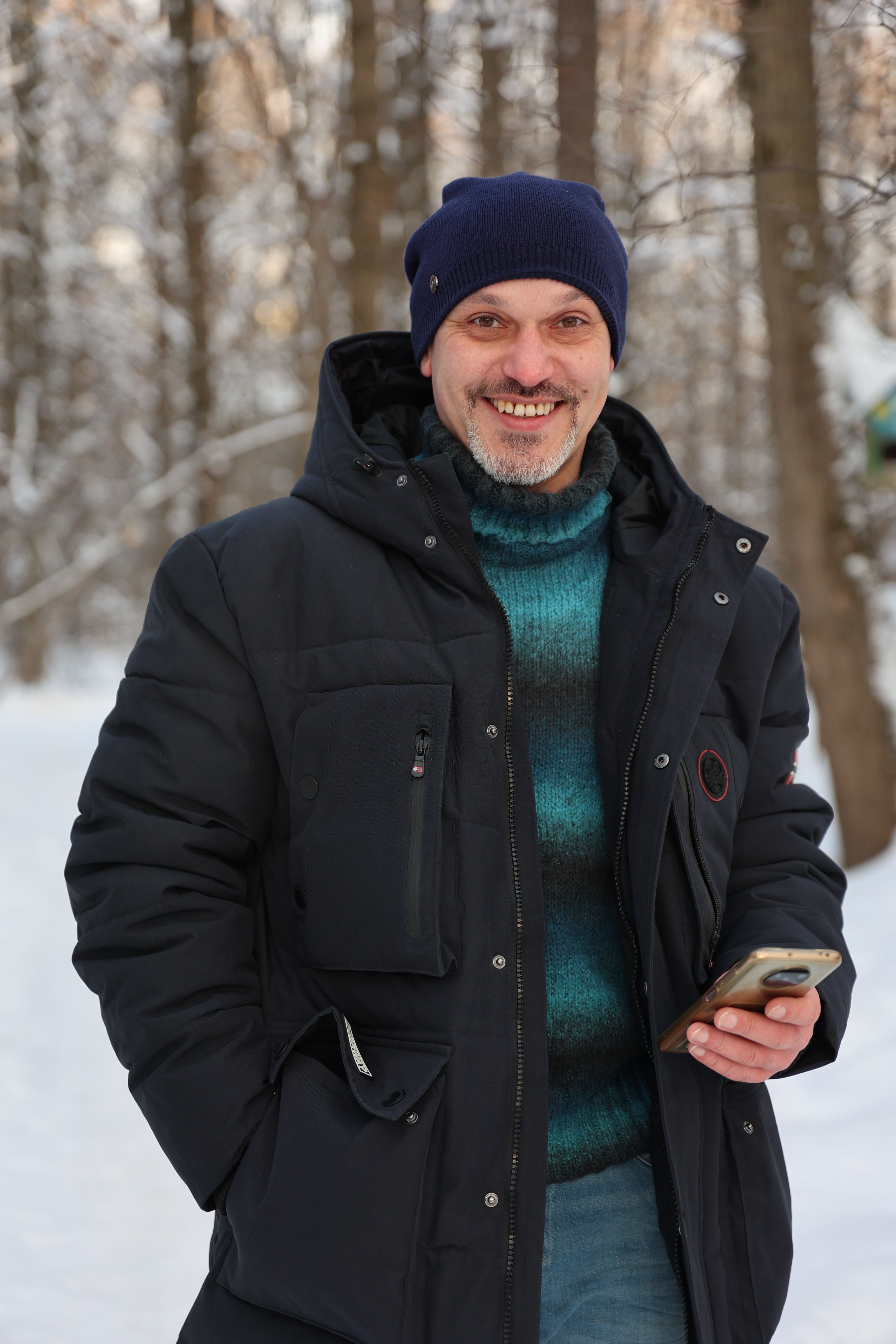 Man standing in snowy forest holding smartphone and smiling, wearing winter clothes.