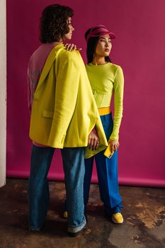Two models posing in vibrant attire against a bold pink backdrop.