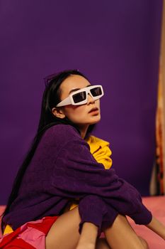 Fashionable young woman in sunglasses poses against vibrant purple studio backdrop, embodying modern style.