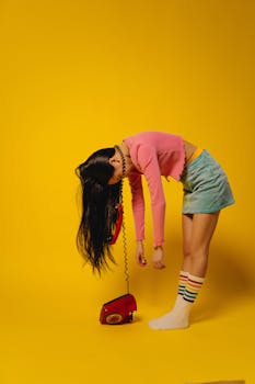 Teenage girl in colorful outfit holding a red telephone against a vibrant yellow background.