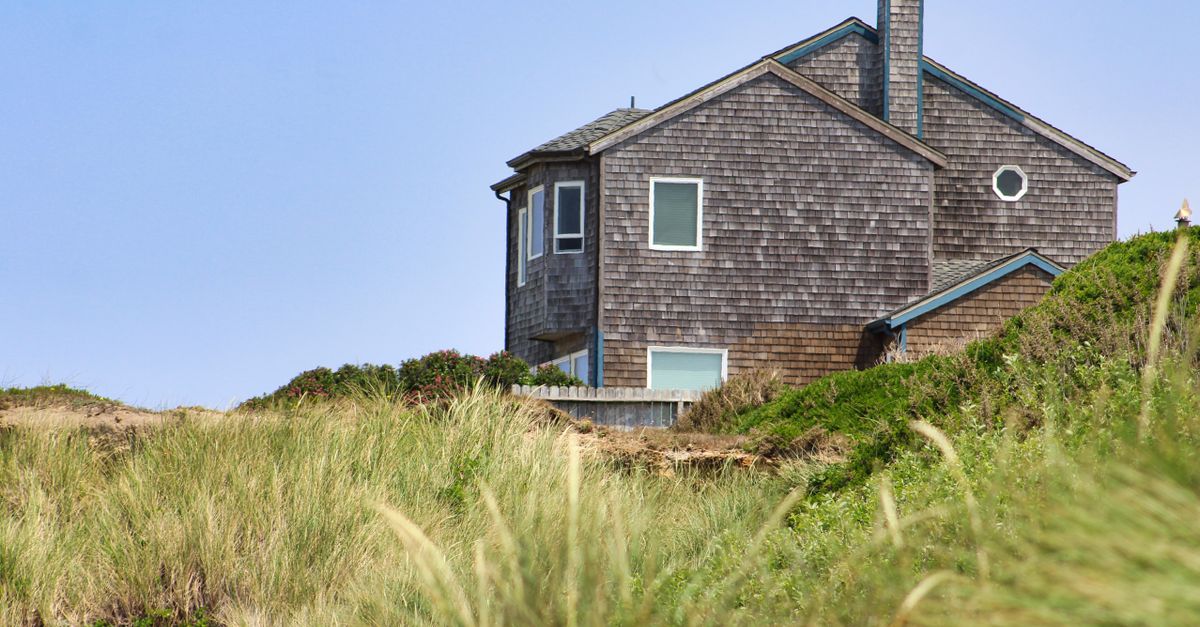 Rustic coastal house nestled on a hill, surrounded by vibrant greenery under clear blue skies.