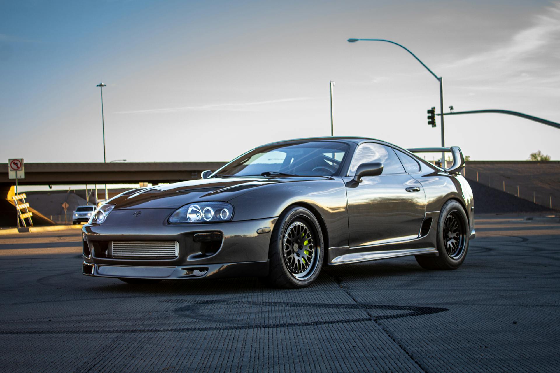 A sleek Toyota Supra sports car is parked on an outdoor asphalt roadway with a sunset backdrop.