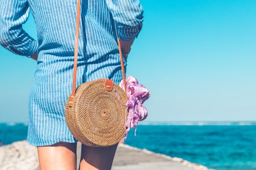 女人穿着蓝色和白色的条纹的裙子与棕色藤藤斜挎包袋附近海洋