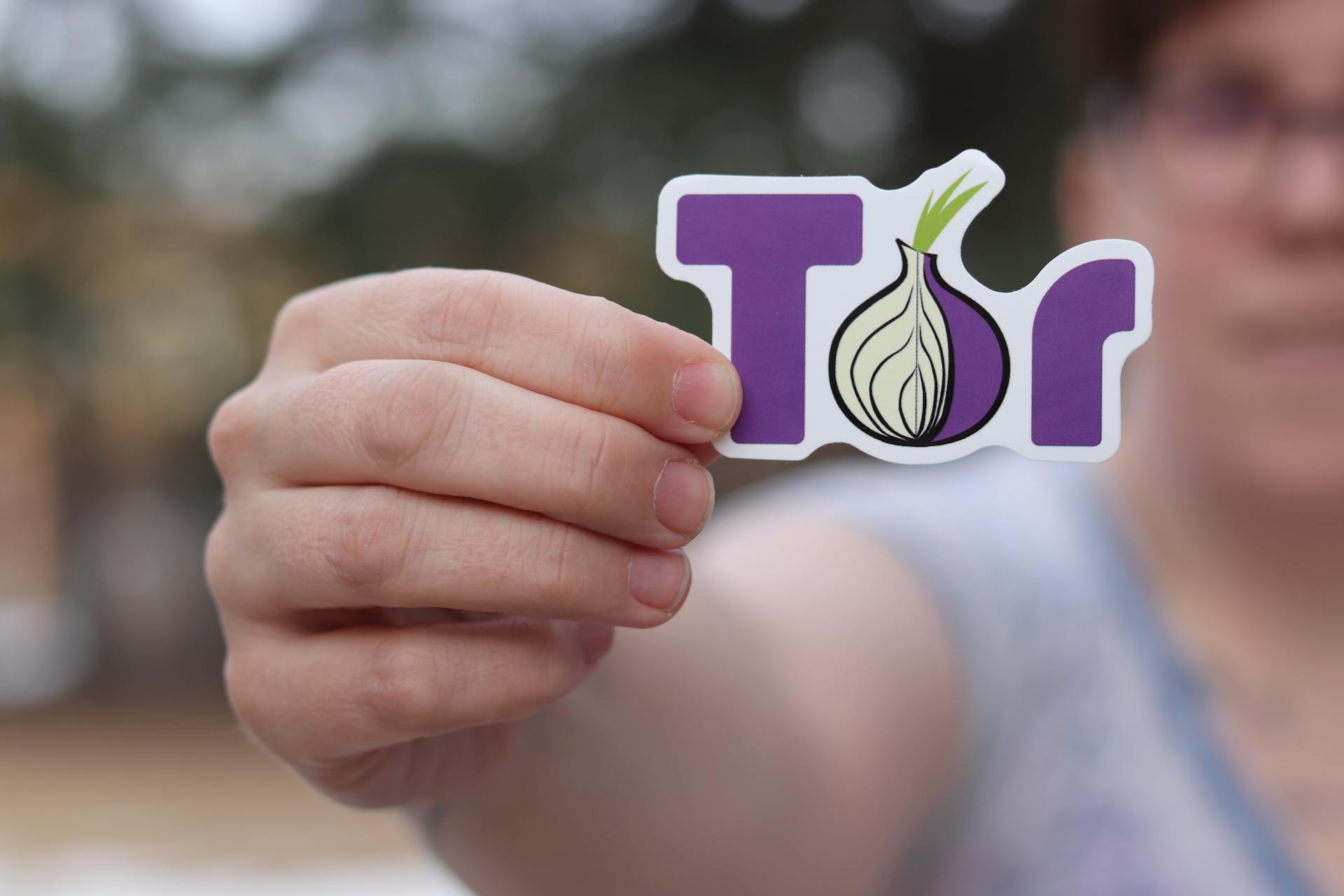 A close-up of a person holding a Tor browser logo sticker with a blurred outdoor background.