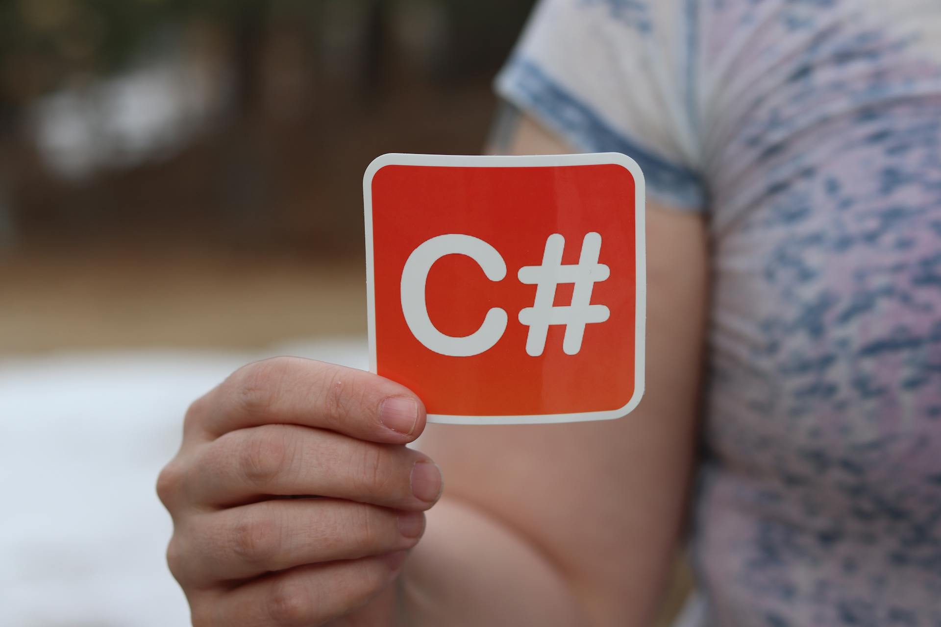 Close-up of a hand holding a C# sticker representing computer programming outdoors.