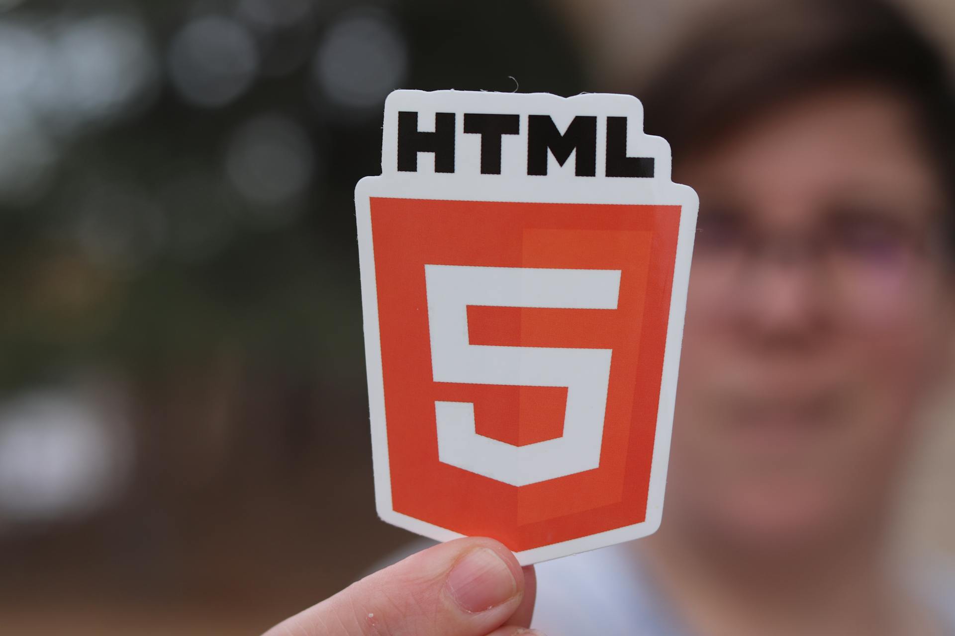 A close-up of an HTML5 logo sticker held by a person with a blurred background, highlighting web development.