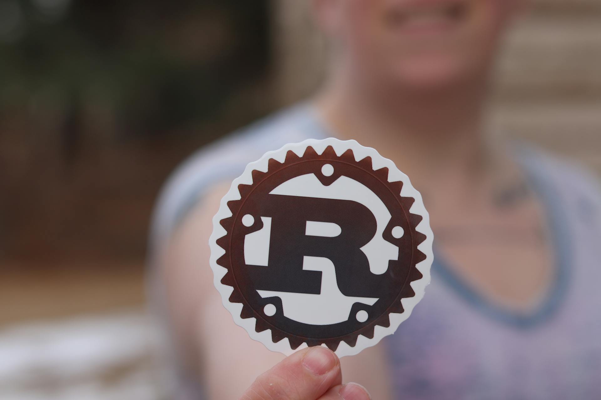 Person holding a Rust programming language logo sticker with blurred background.