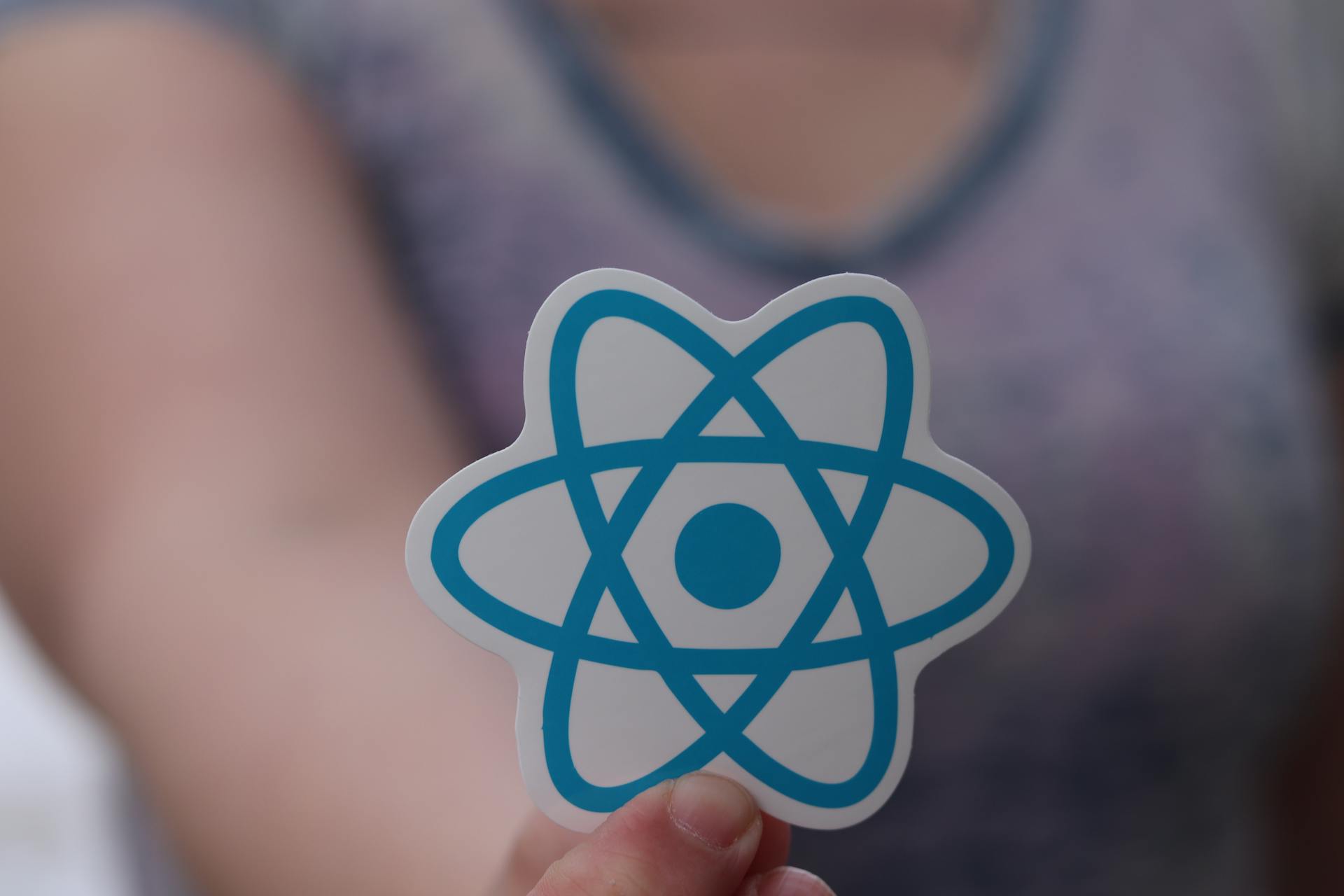 A person holds a sticker featuring the React logo, commonly used in web development.