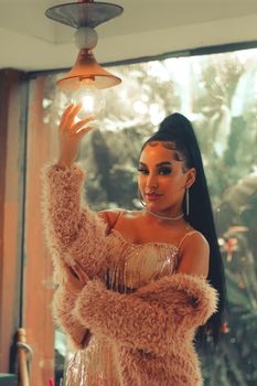 Woman in a fur coat poses indoors holding a light bulb with elegant style.