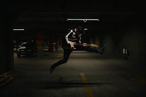 Person in Black long Sleeve Shirt Jumping on Parking Lot and Holding an Electric Guitar