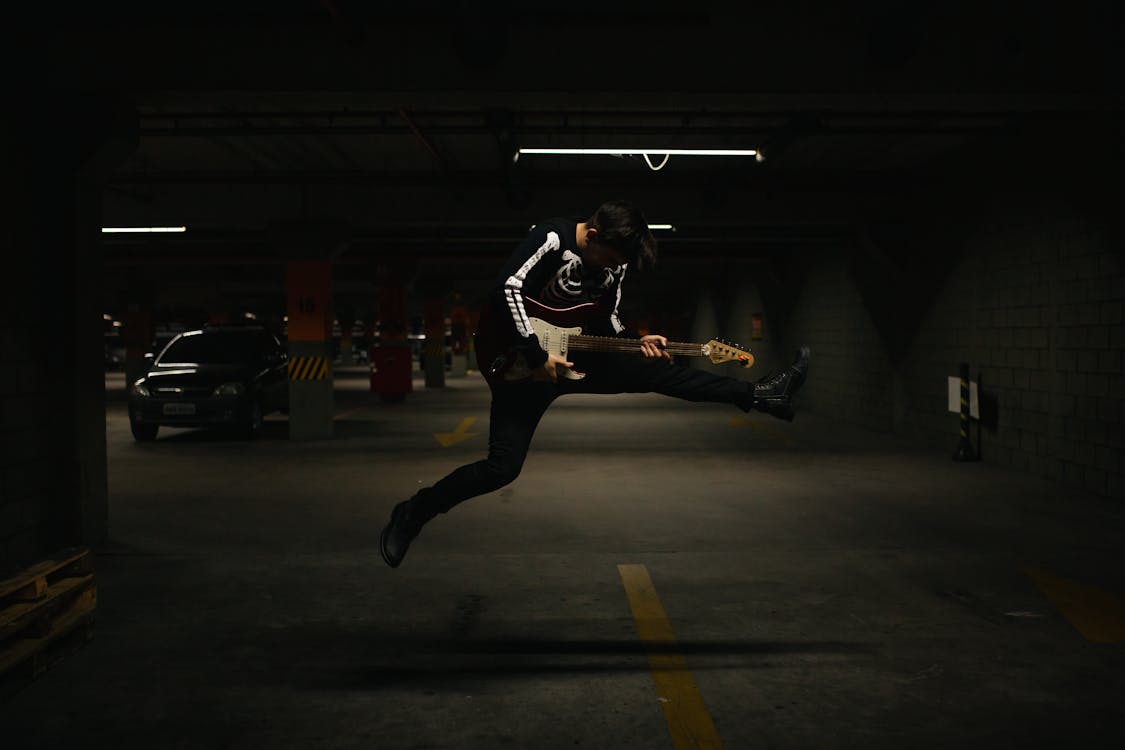 Person in Black long Sleeve Shirt Jumping on Parking Lot and Holding an Electric Guitar